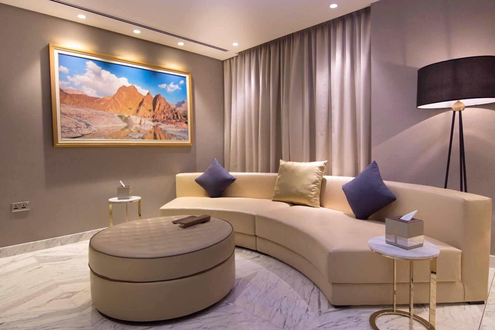 Un salon moderne avec un canapé aux lignes courbes, un pouf rond, un tableau représentant un paysage désertique, une lampe sur pied et une petite table d’appoint.