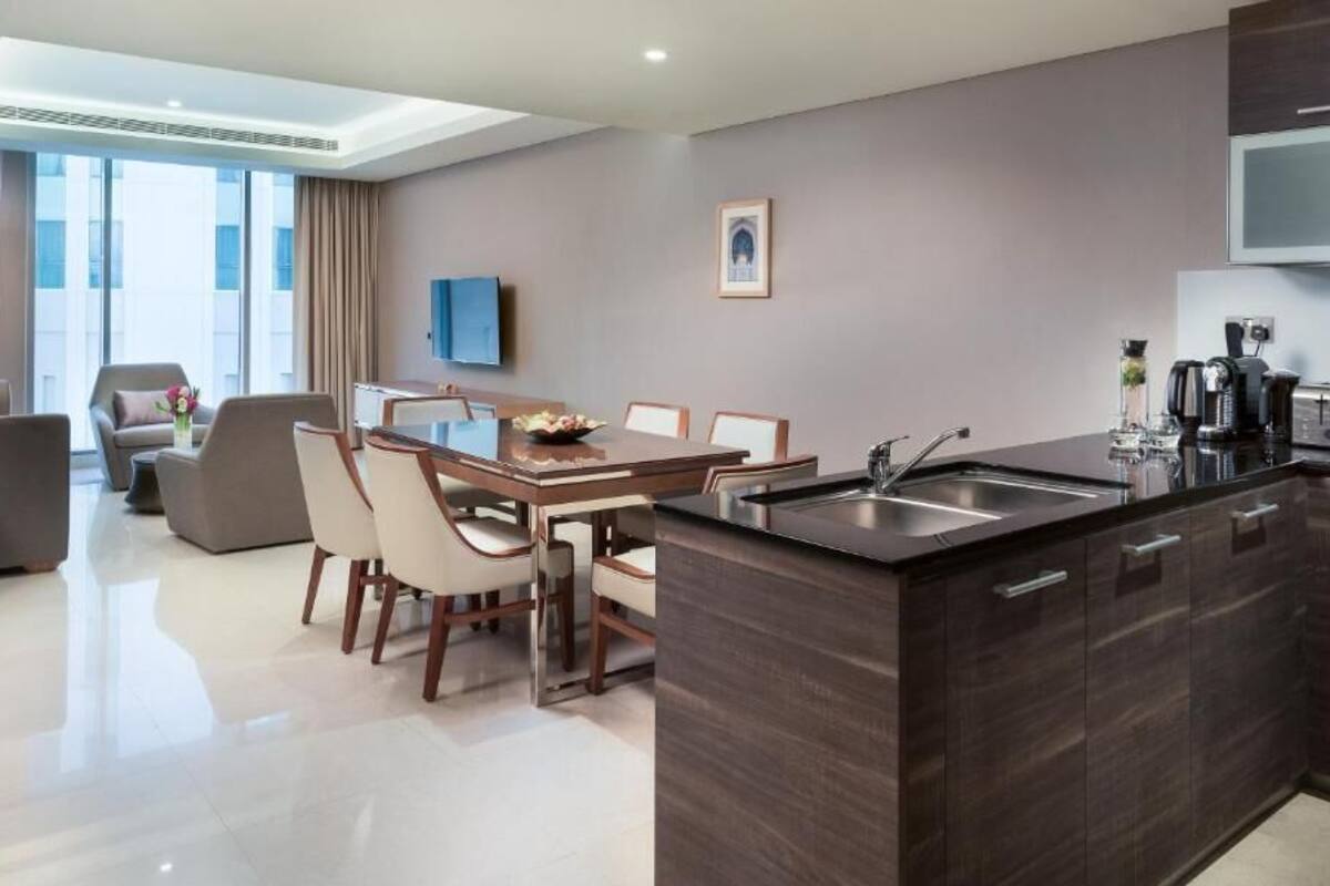 A modern hotel room with a dining area, a kitchen with a sink, and a flat-screen TV.