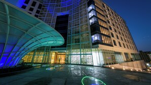 A modern hotel with a curved glass facade, multiple floors, and illuminated signage. - Grand Millennium Muscat Apartments and Suites connected to Mall (Muscat)