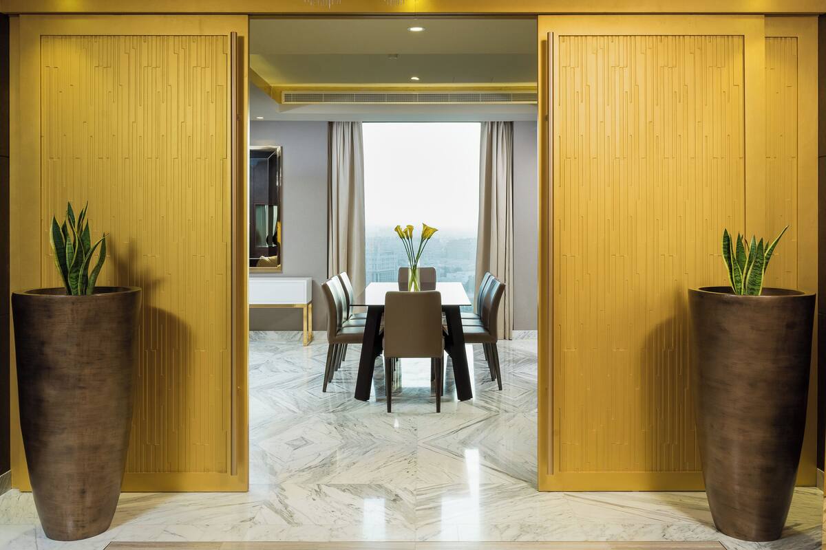 A hotel room with a dining area, large potted plants, and a view through a window.