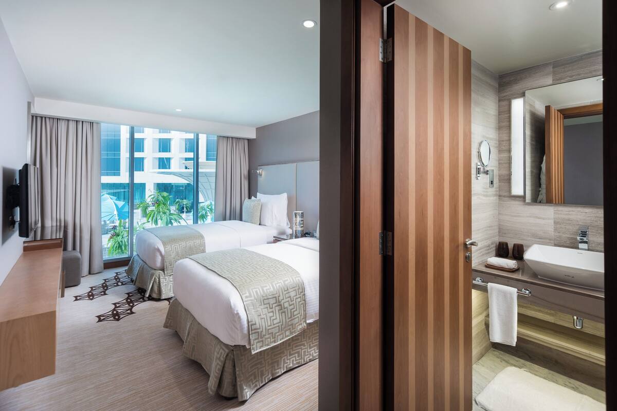 A hotel room with a large window, a bed, a bedside table, a bathroom with a sink and mirror, and a view of a building outside.