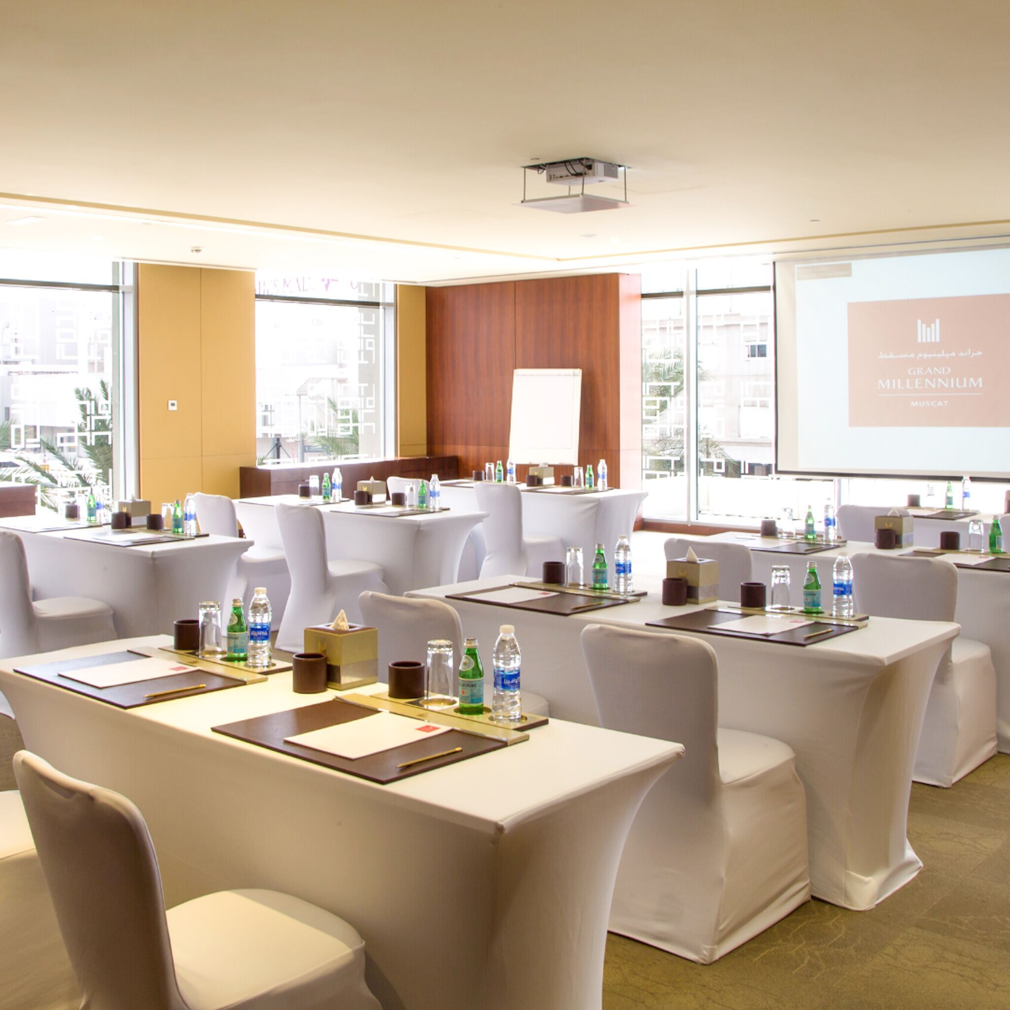 Un salón de conferencias con mesas, sillas y una pantalla de proyector que muestra 'Grand Millennium Muscat'.