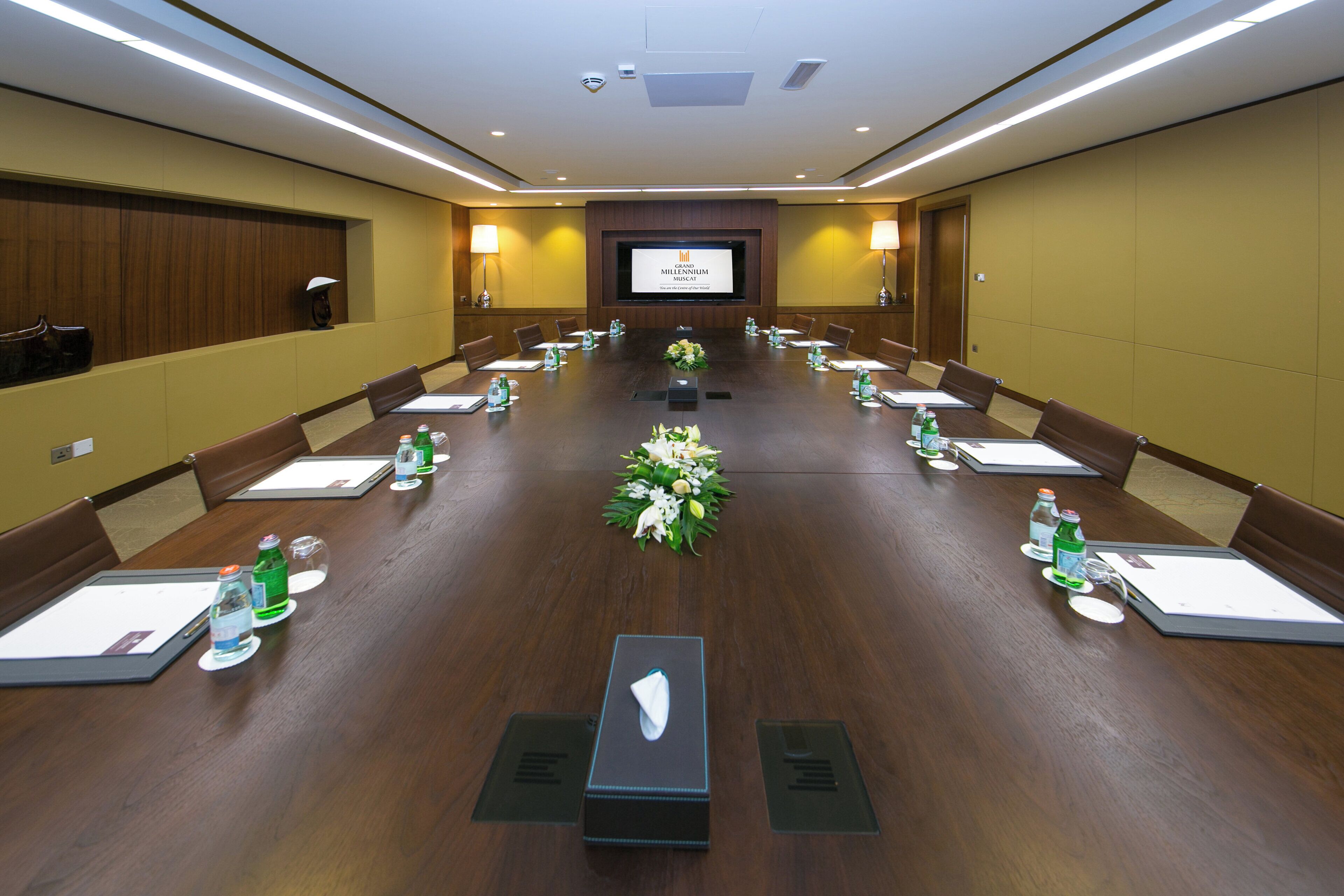 A conference room with a long table, chairs, a projector screen, and a floral centerpiece.