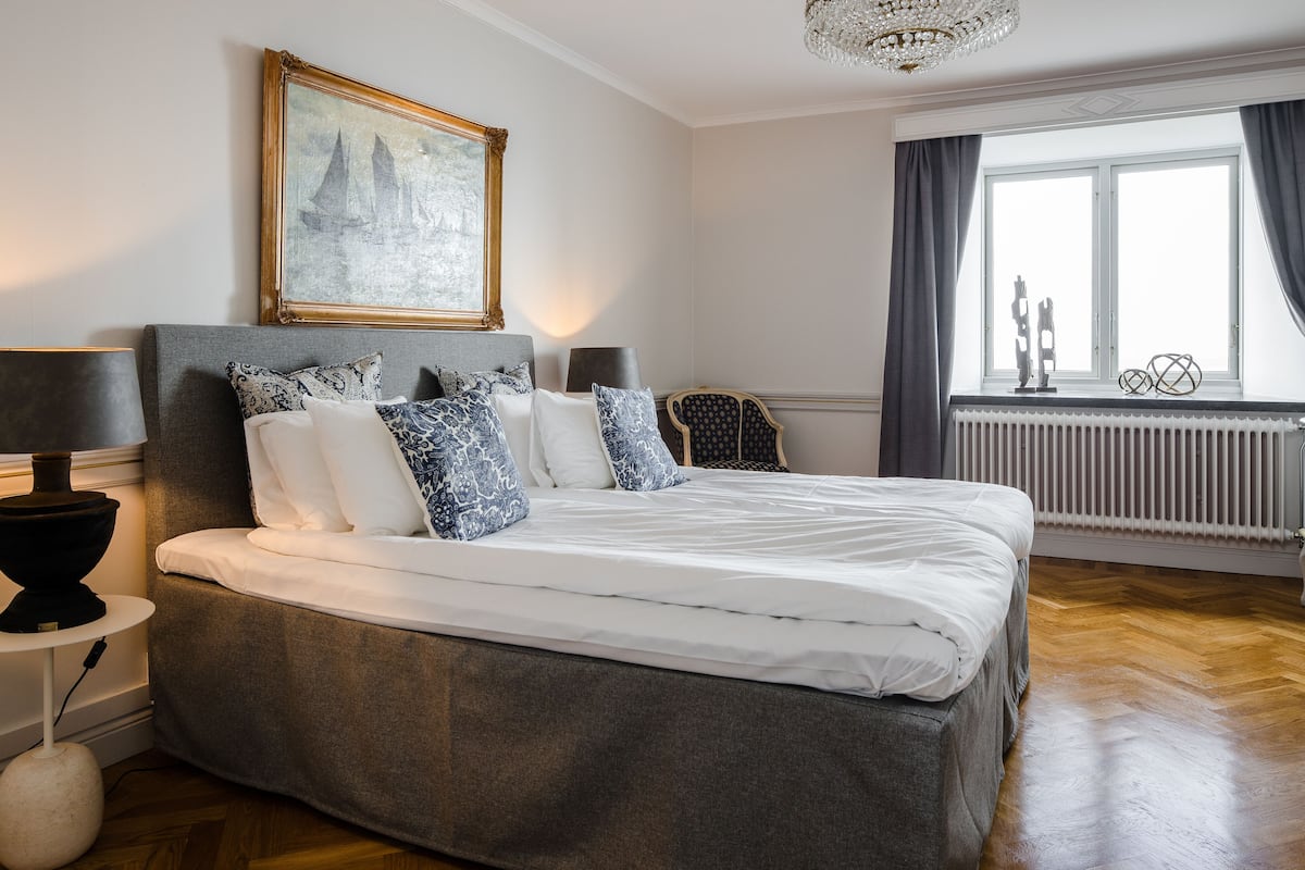 A neatly made bed with a grey headboard, a framed painting above it, and a window with curtains to the right.