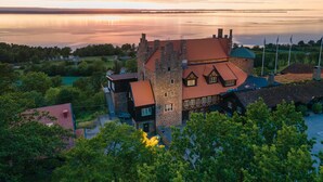 A historic building with a stone tower and red-tiled roofs, surrounded by lush greenery and a calm lake. - Hotel Gyllene Uttern (Gränna)