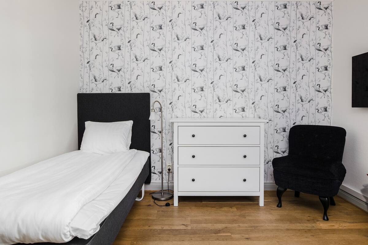 A bedroom with a bed, a white dresser, a black chair, and a wall with a bird pattern.