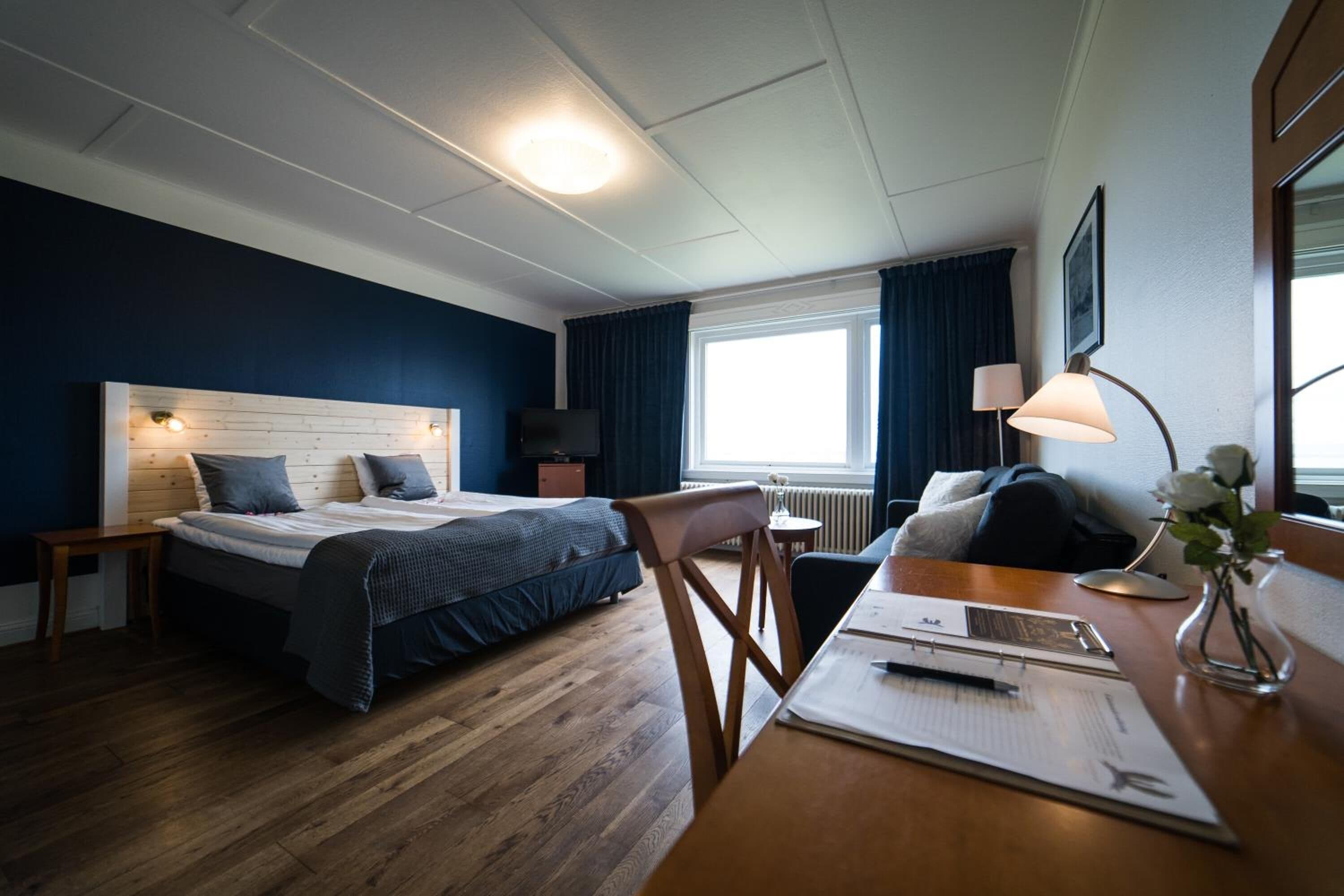 A hotel room with a bed, a desk with a chair, a lamp, and a window with curtains.