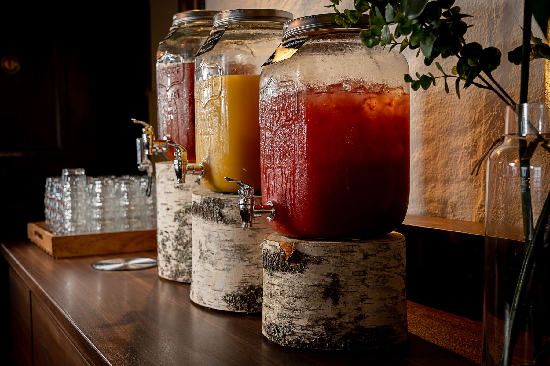 A bar area with multiple drink dispensers on birch logs, glasses stacked on a wooden surface, and a plant in the background.