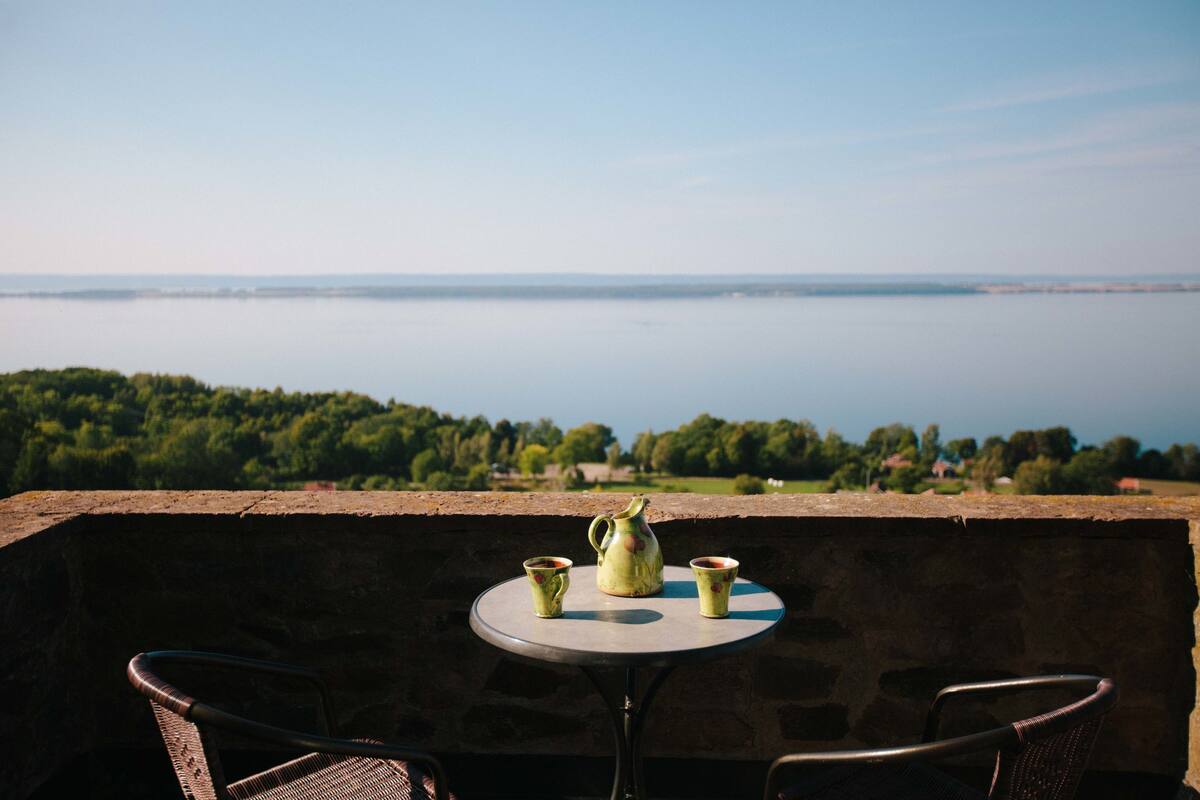 A balcony with a table and two chairs, overlooking a body of water and greenery.