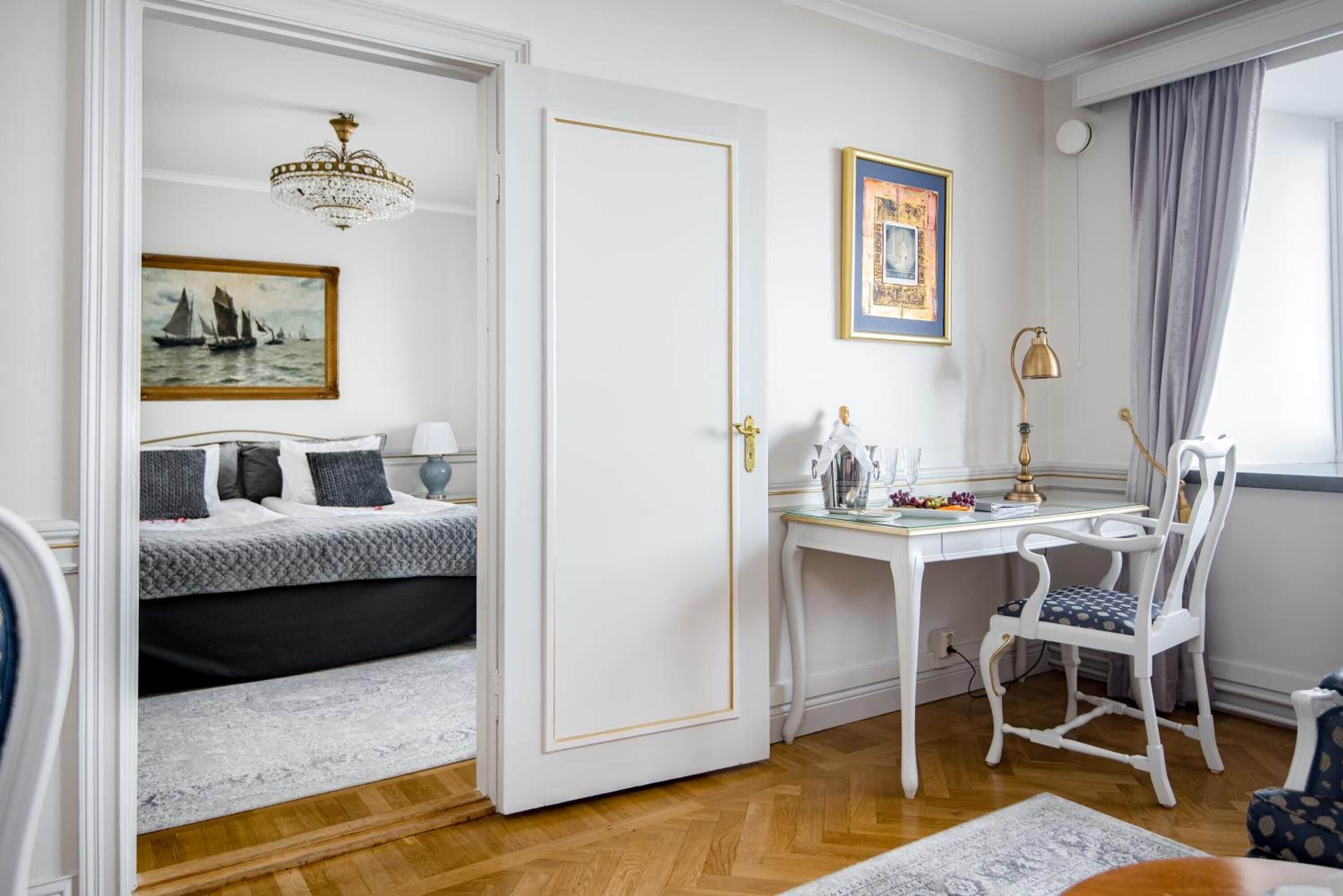Una habitación con cama, escritorio, silla, candelabro y un cuadro en la pared.