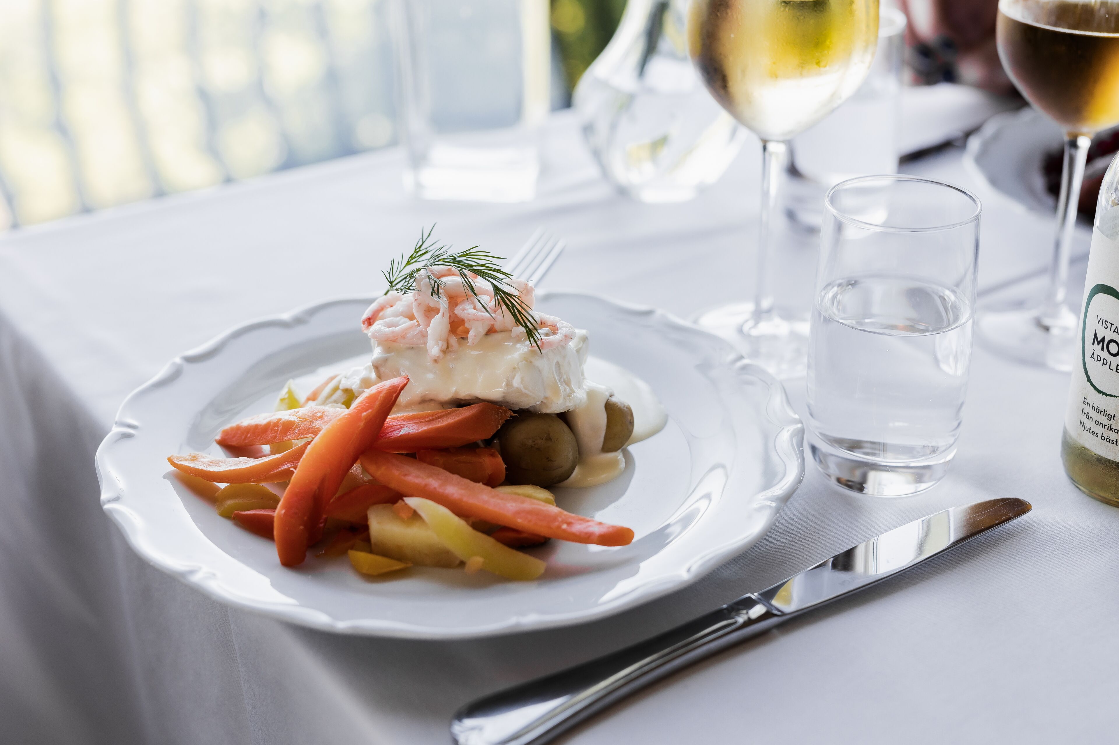 A plate of food with a fish dish, carrots, and potatoes, accompanied by a glass of water and a bottle of wine on a table.