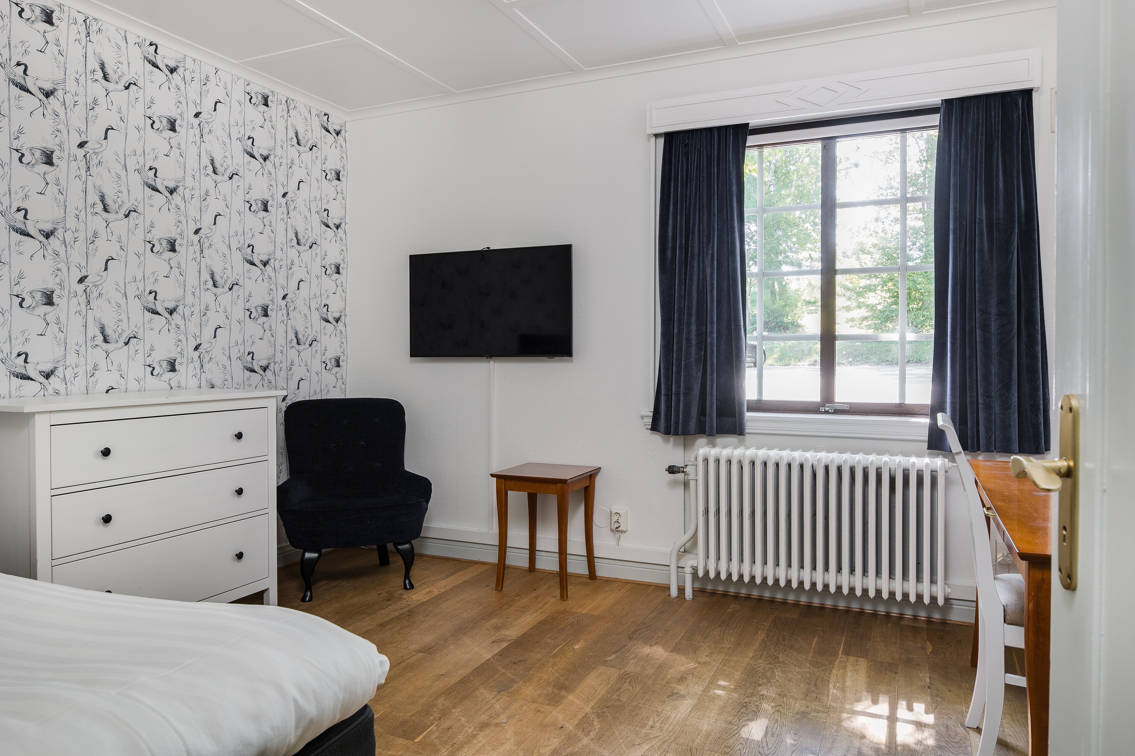 Une pièce comprenant un lit, une commode, une chaise noire, une petite table en bois, un radiateur et une fenêtre avec des rideaux.