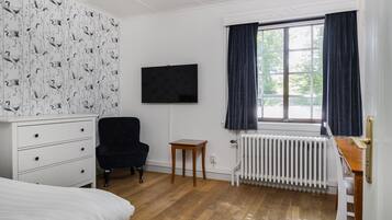 A room with a bed, a dresser, a black chair, a small wooden table, a radiator, and a window with curtains.
