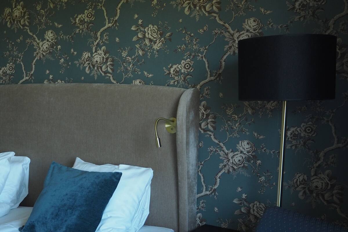 A hotel room with a bed, two pillows, a nightstand, a lamp, and floral wallpaper.