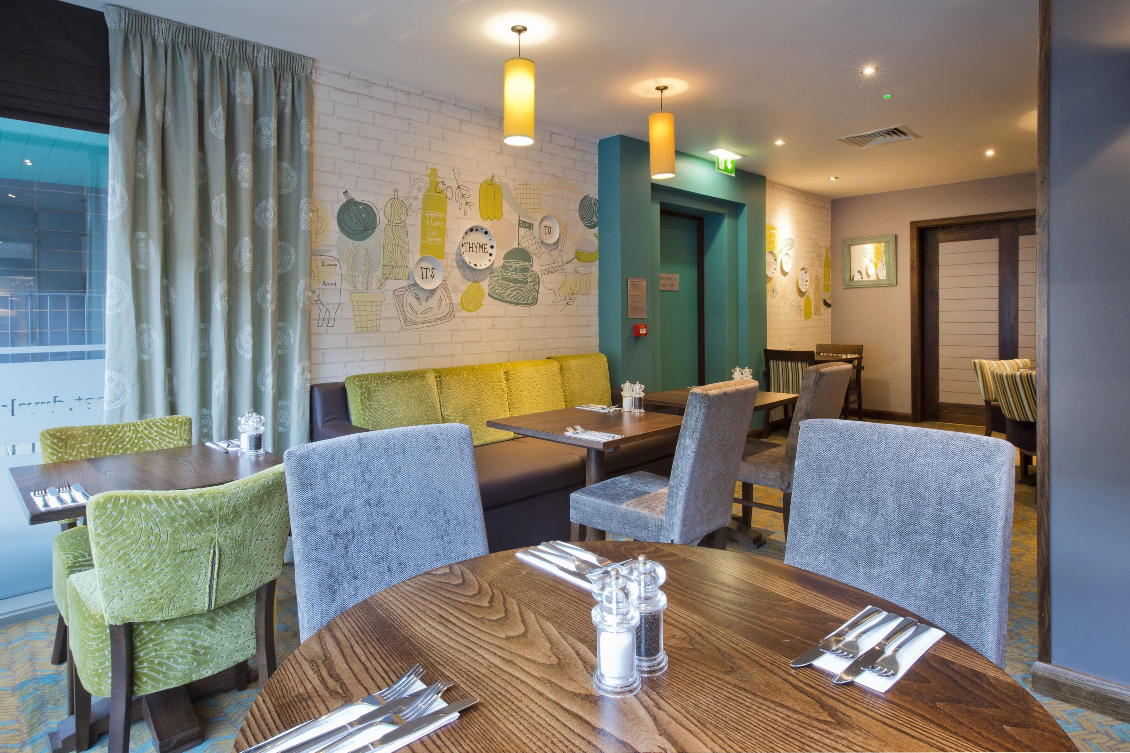 Comedor con mesas de madera, sillas tapizadas de verde y una pared de ladrillo decorada con obras de arte.