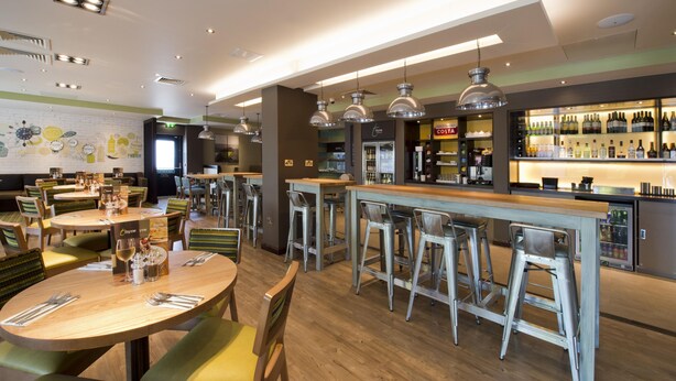 Un restaurant avec des tables rondes, des tabourets de bar, des luminaires suspendus, un coin bar avec des bouteilles et un mur orné d’une horloge et d’un menu.