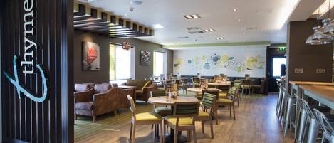 A modern restaurant with wooden flooring, round tables, and chairs. There is a bar area with stools.