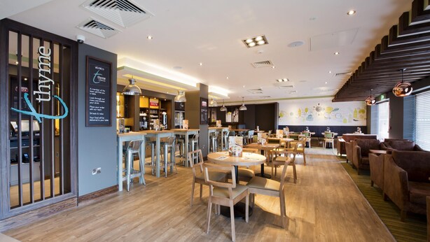 A modern restaurant interior with wooden flooring, round tables, and bar seating. - Premier Inn Honiton (Honiton)