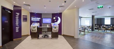 Zona de recepción de un hotel con mostrador de registro, pantalla digital y un cartel de 'Premier Inn'.