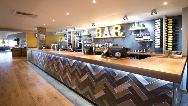 Een bar met een visgraatvloer, een toonbank met kassa en planken vol flessen en glazen.