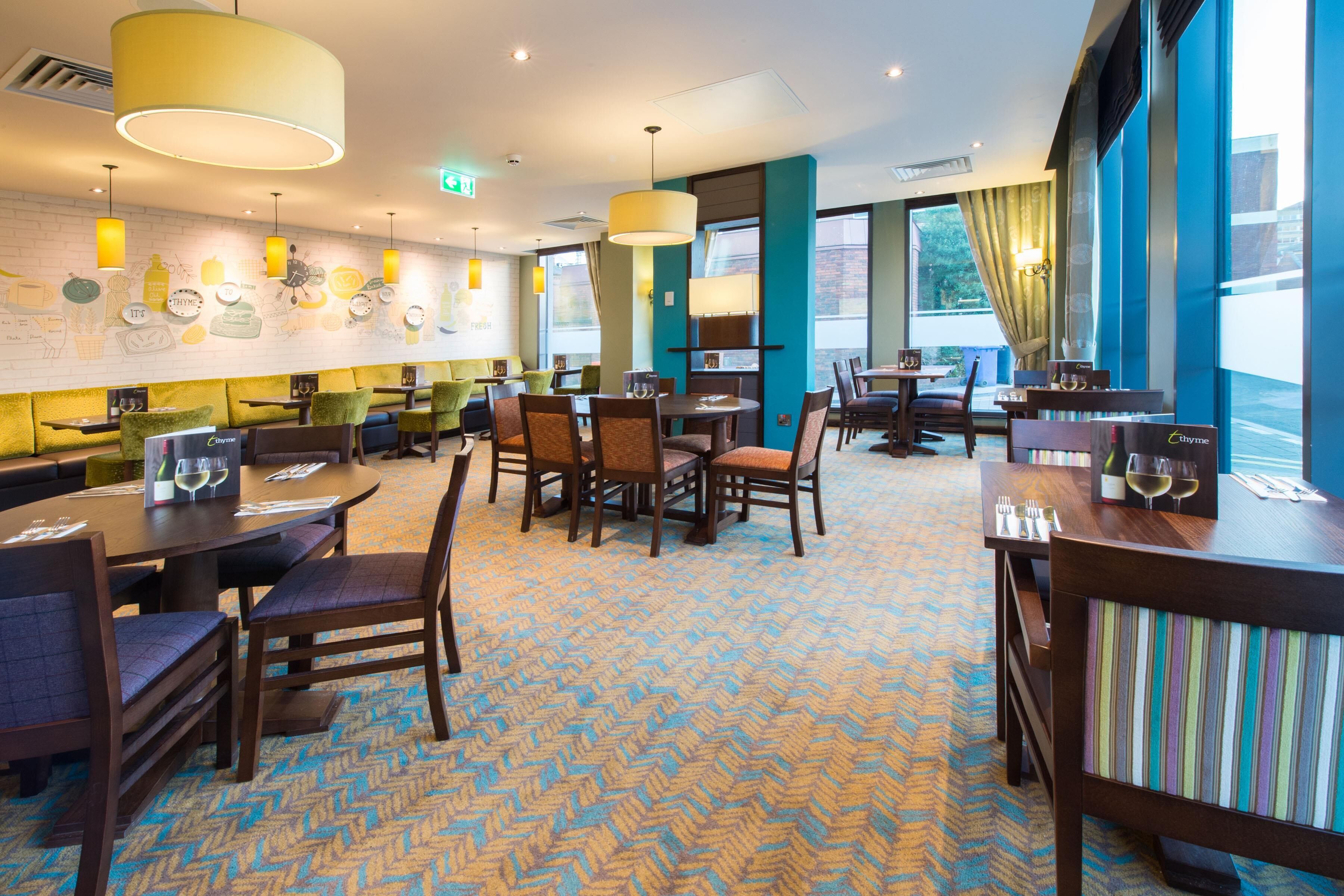 En hotelrestaurant med flere borde og stole, store vinduer og et mønstret tæppe.