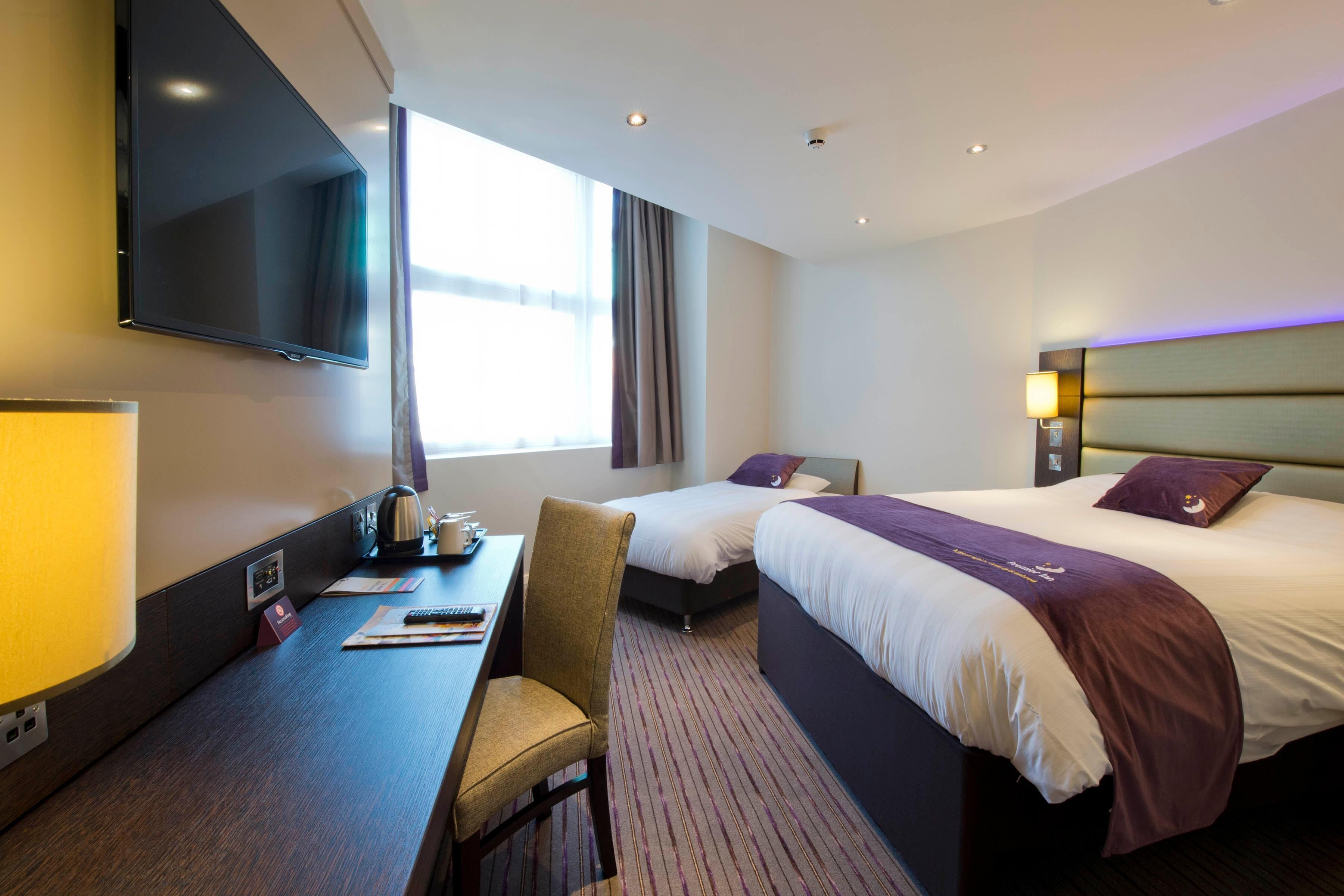 A hotel room with a bed, a desk with a chair, a TV mounted on the wall, and a window with curtains.