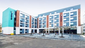 Et moderne hotel med flere etager, med facader af både mursten og glas, udendørs siddepladser og en parkeringsplads.