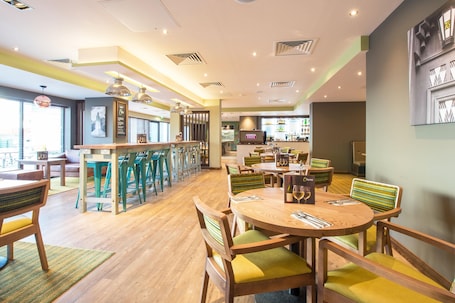 A restaurant with a bar area, wooden tables, and chairs with green cushions.