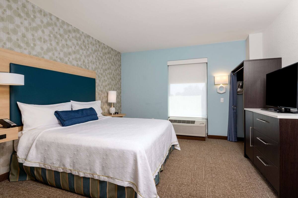 Une chambre d’hôtel équipée d’un lit, d’une table de chevet, d’une lampe, d’une télévision et d’une fenêtre avec des stores.