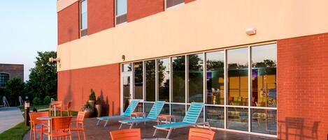 An outdoor patio area with orange chairs and tables, a brick building with large windows, and a grassy area.