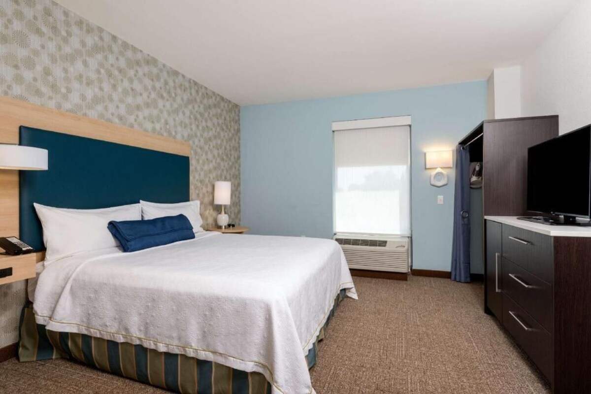 Habitación de hotel con cama, mesita de noche, lámpara, televisor, armario y ventana con persianas.