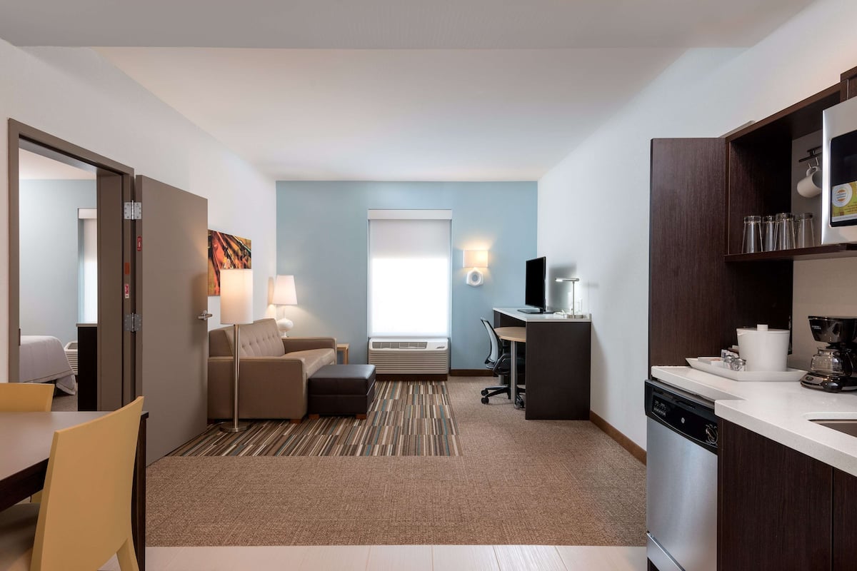 Une chambre d’hôtel moderne dotée d’une kitchenette, d’un canapé, d’un coin repas et d’une grande fenêtre laissant entrer la lumière naturelle.