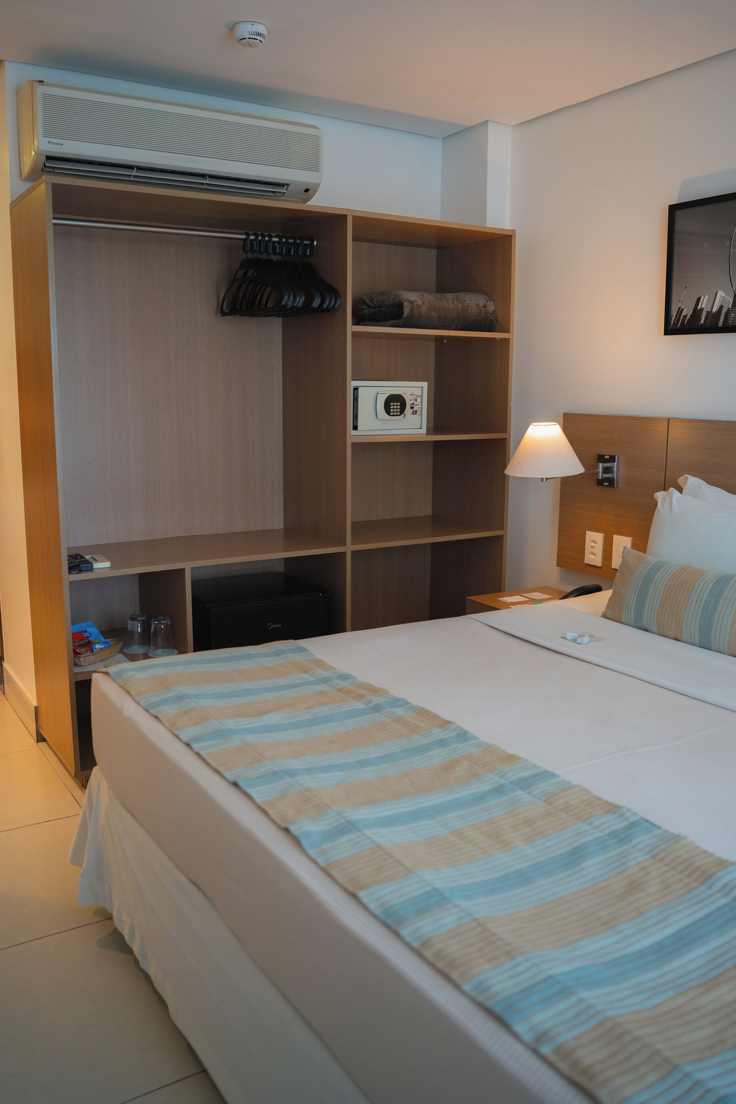 Quarto de hotel com cama, guarda-roupa, secretária, abajur e ar condicionado.