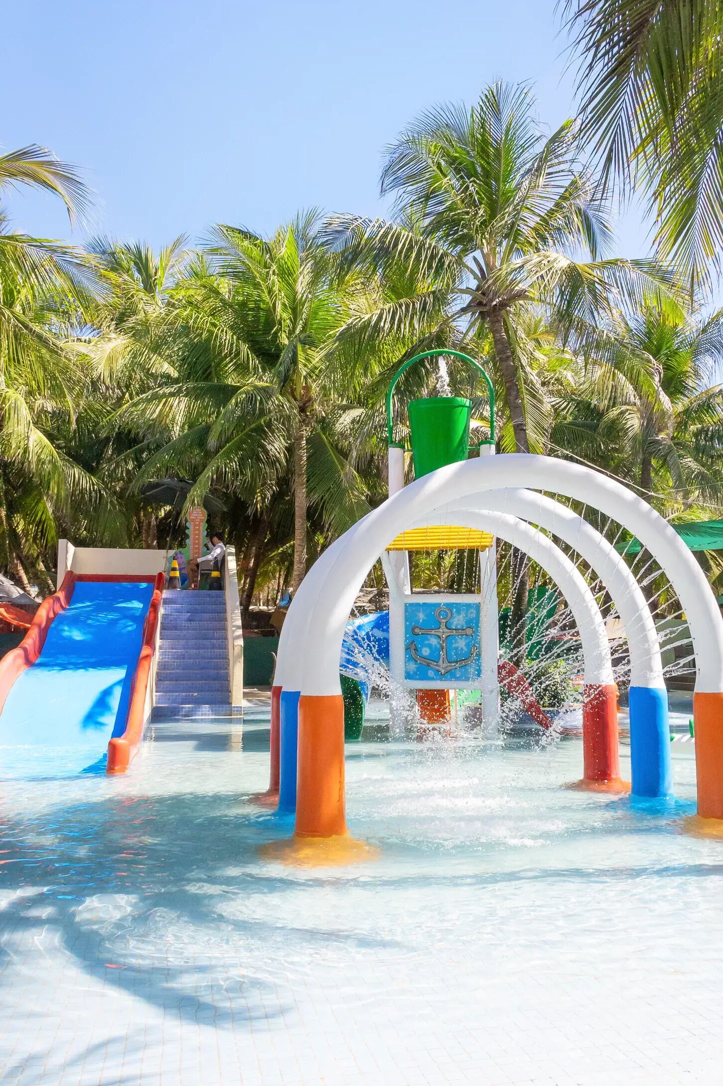 Ein Wasserpark mit einer blauen Rutsche, einem Wasserspielbereich und Palmen im Hintergrund.