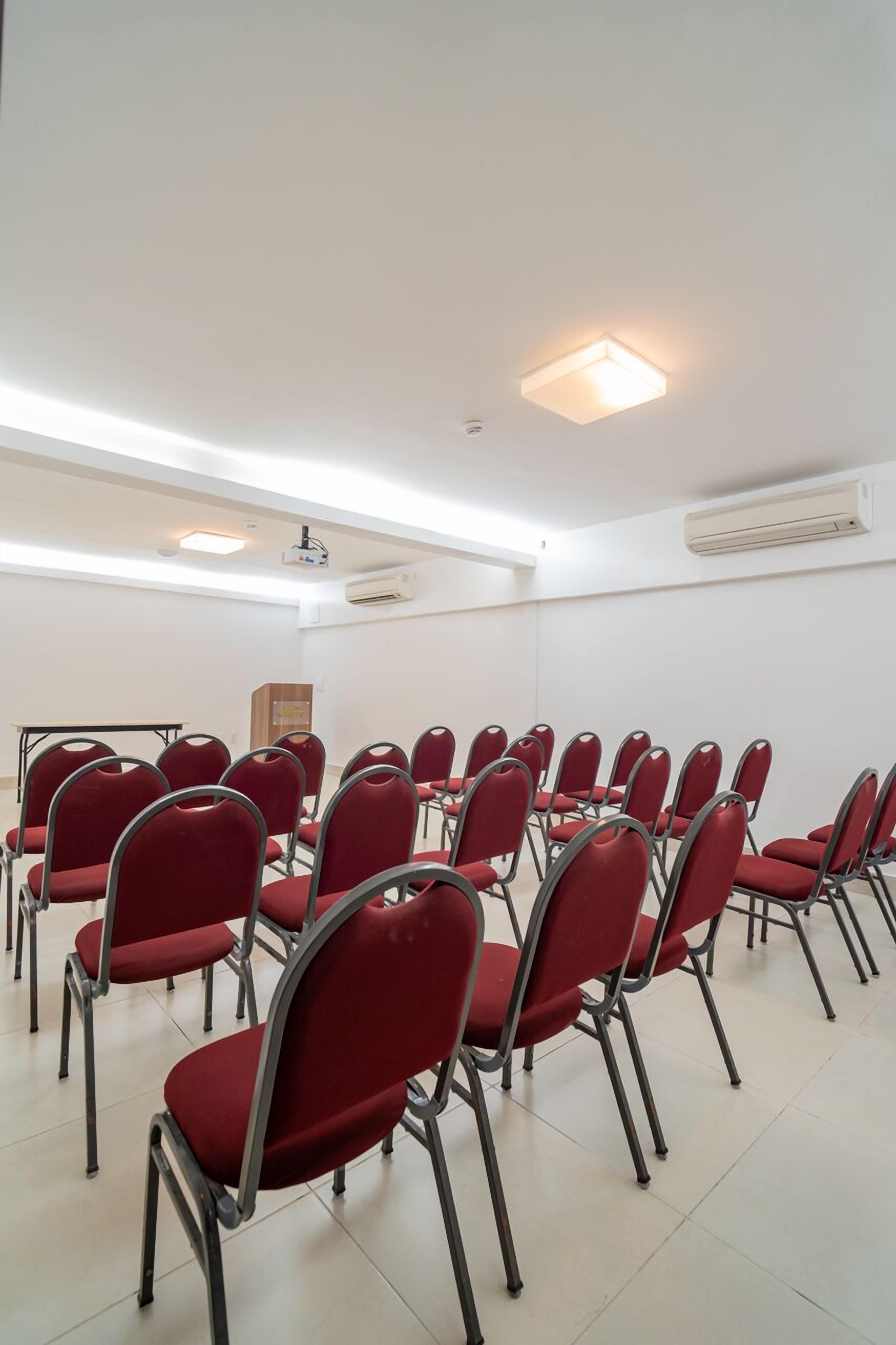 A conference room with rows of red chairs, a projector, and an air conditioning unit on the wall.