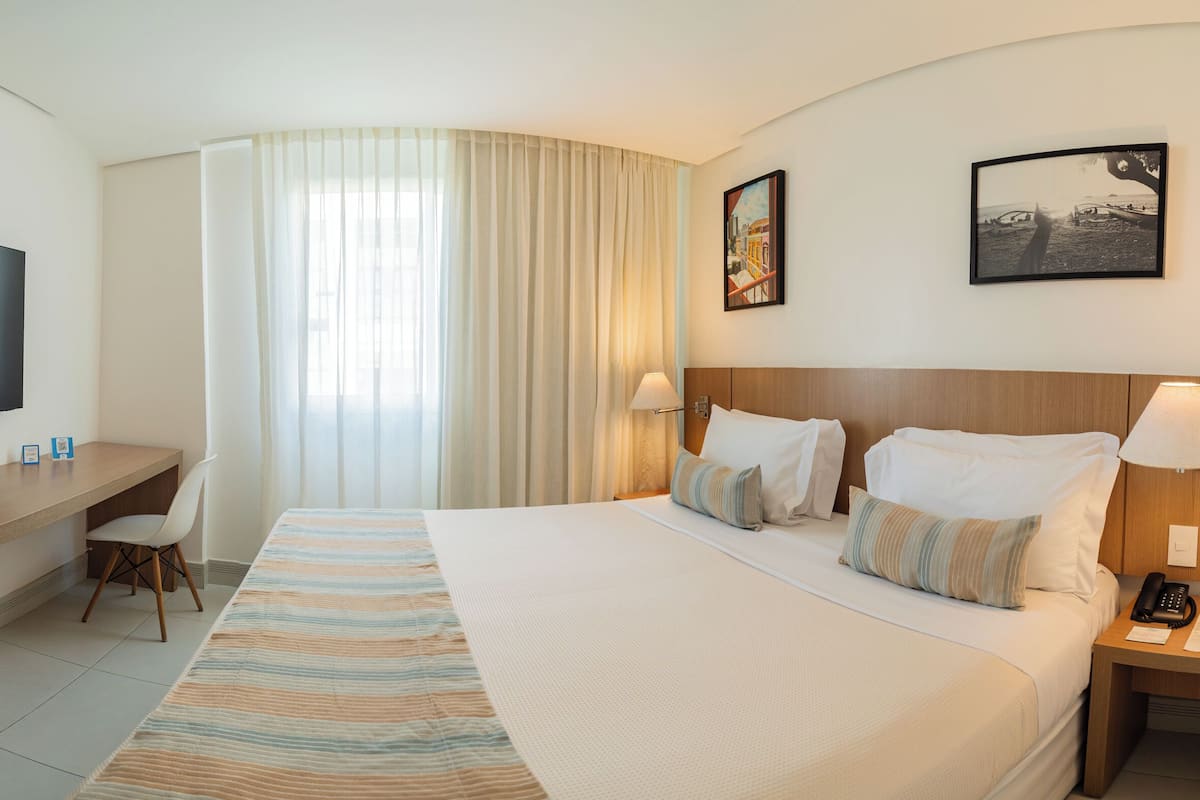 A hotel room with a bed, a desk, a chair, a TV, and a lamp.