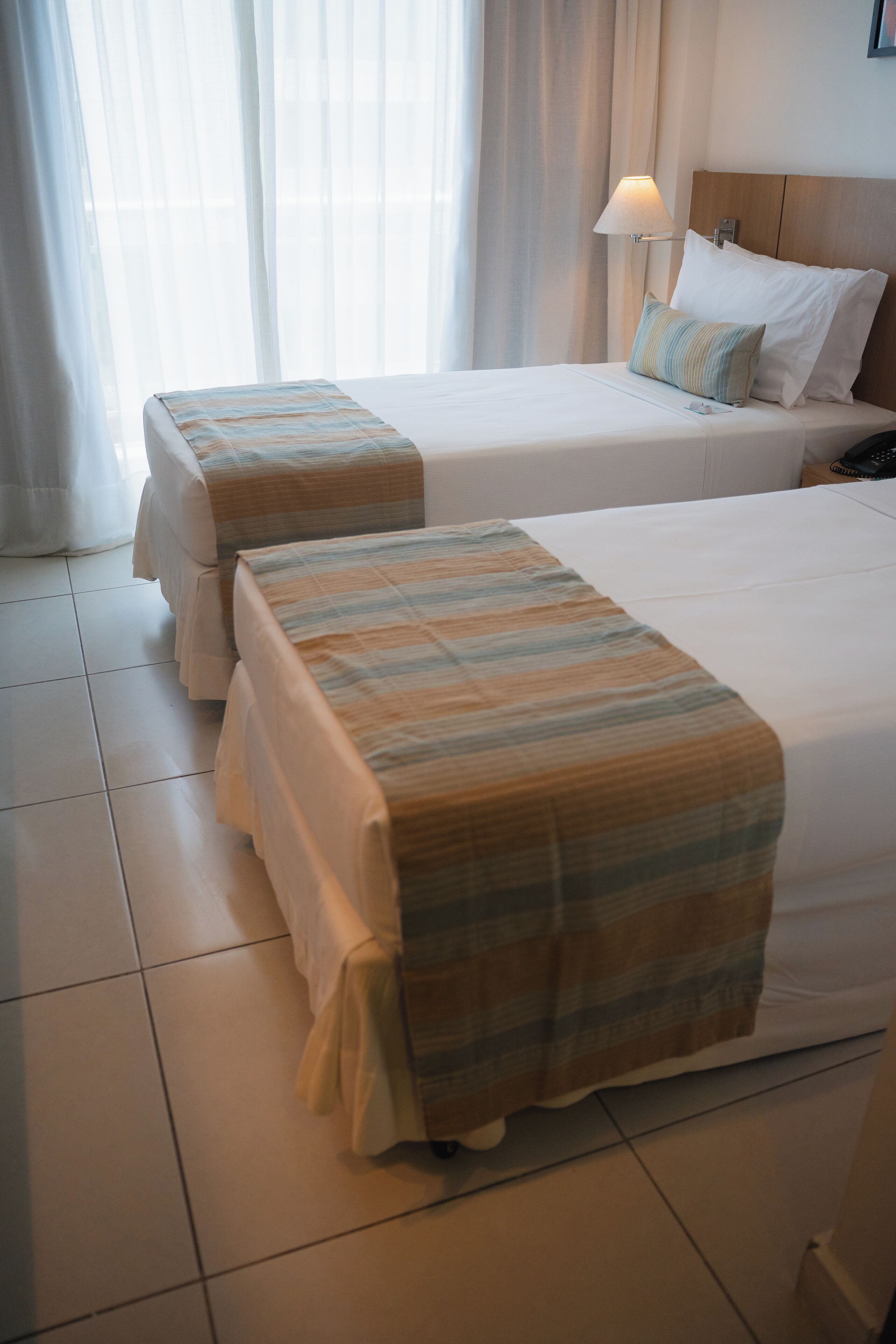 A hotel room with two beds, a bedside table, a lamp, and a window with curtains.