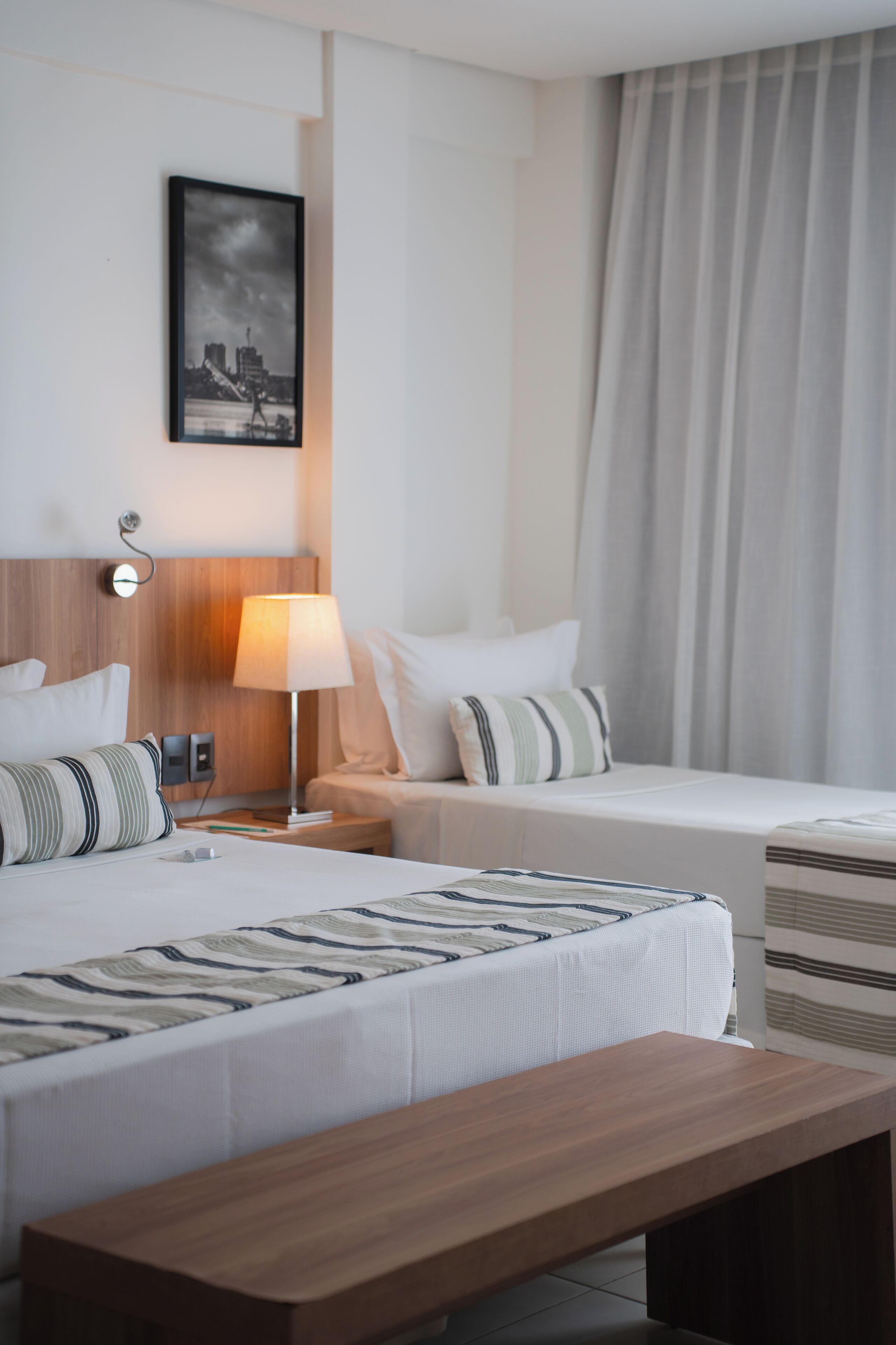 A hotel room with two beds, a wooden bench, a bedside table with a lamp, a framed picture on the wall, and a window with curtains.