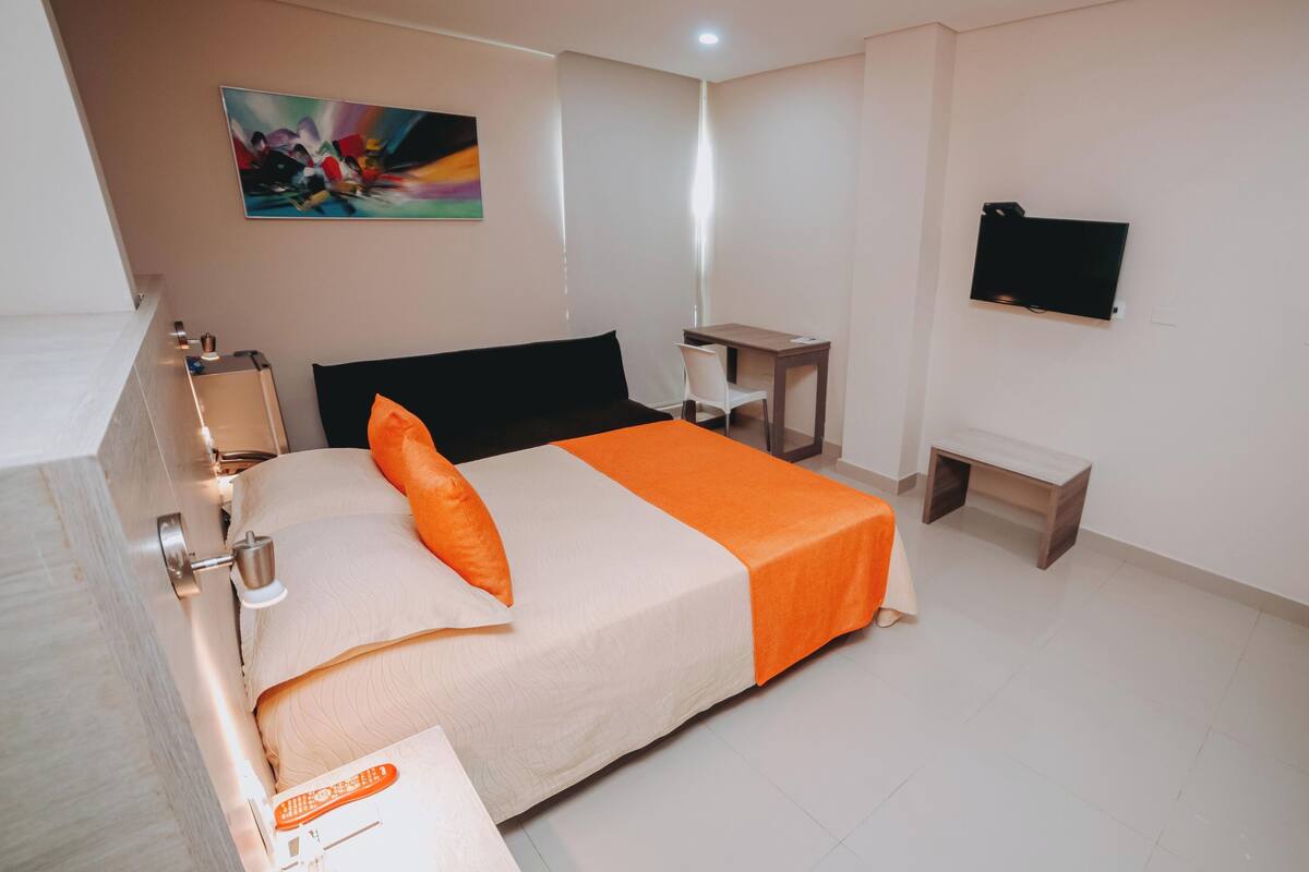 A hotel room with a bed, a desk, a chair, a TV, and a painting on the wall.