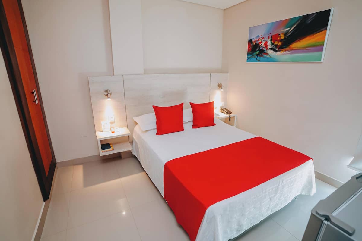A hotel room with a large bed, white bedding, red pillows, a nightstand with a phone, and a painting on the wall.