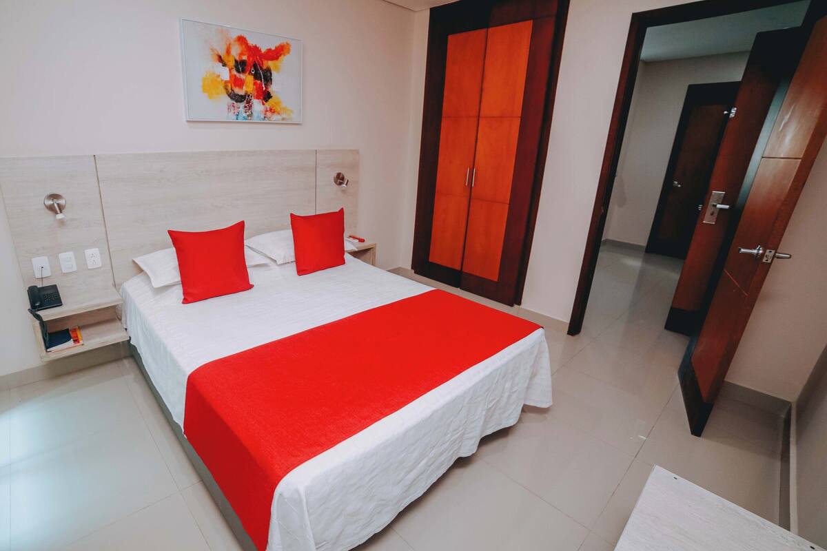 A hotel room with a bed, a nightstand, a bedside table, a red blanket, a red pillow, a wall-mounted painting, and a wardrobe.