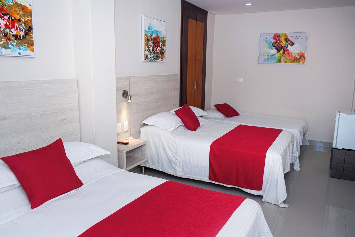 A hotel room with three beds, each with white bedding and red pillows. There are two paintings on the walls, a nightstand with a lamp, and a small desk with a computer.