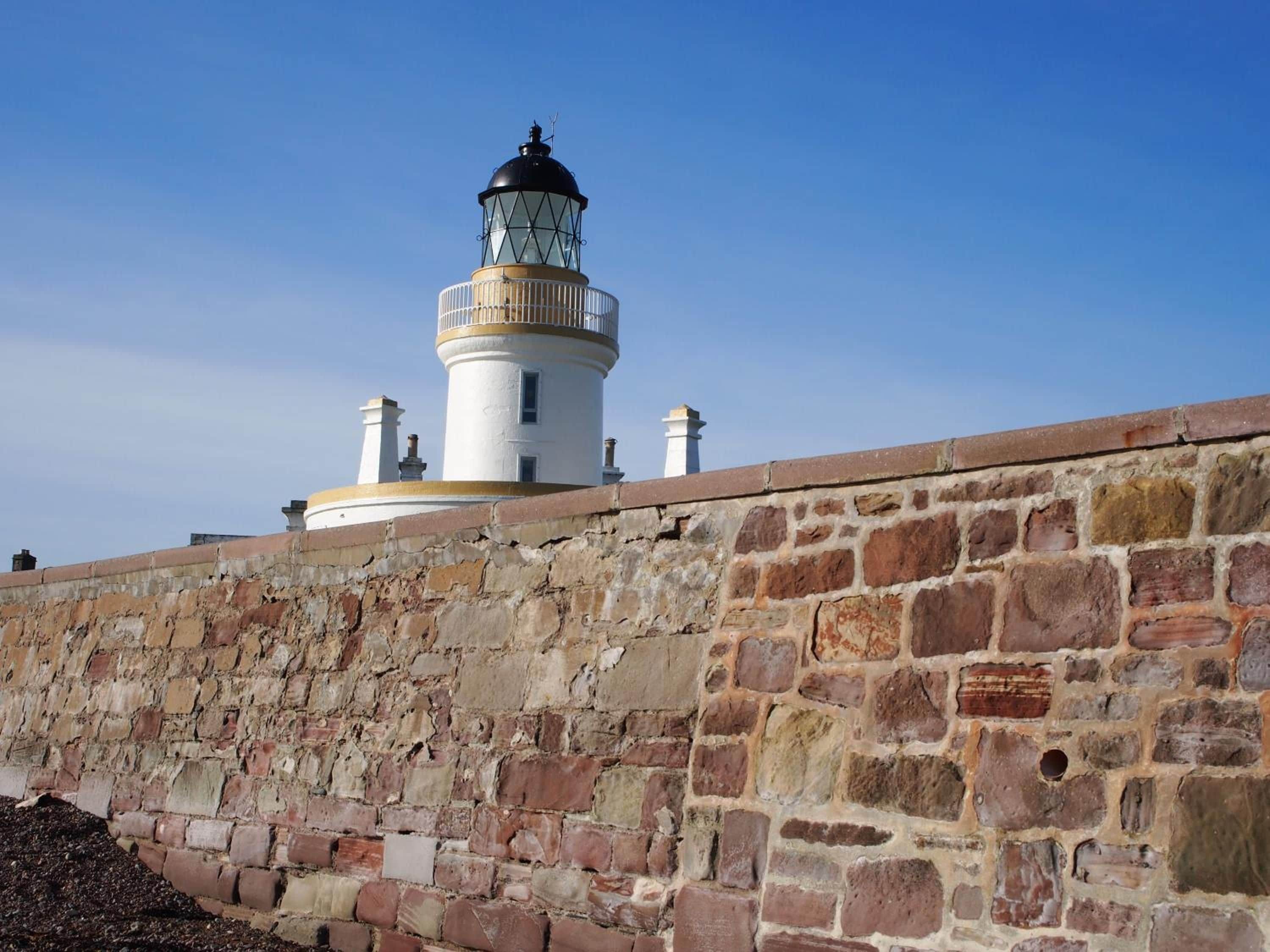 A lighthouse with a white cylindrical structure and a black top, situated next to a stone wall.