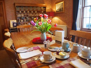 A dining area with a wooden table set for four, a vase of tulips, and a kitchen in the background with various dishes and utensils.