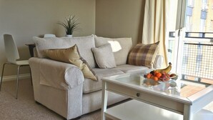 A living room with a beige sofa, a glass coffee table with fruit, a chair, and a plant. - Marine View (Dundee)