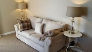 A beige sofa with a plaid throw blanket, two wall-mounted lamps, and a small side table with a lamp. - Marine View (Dundee)