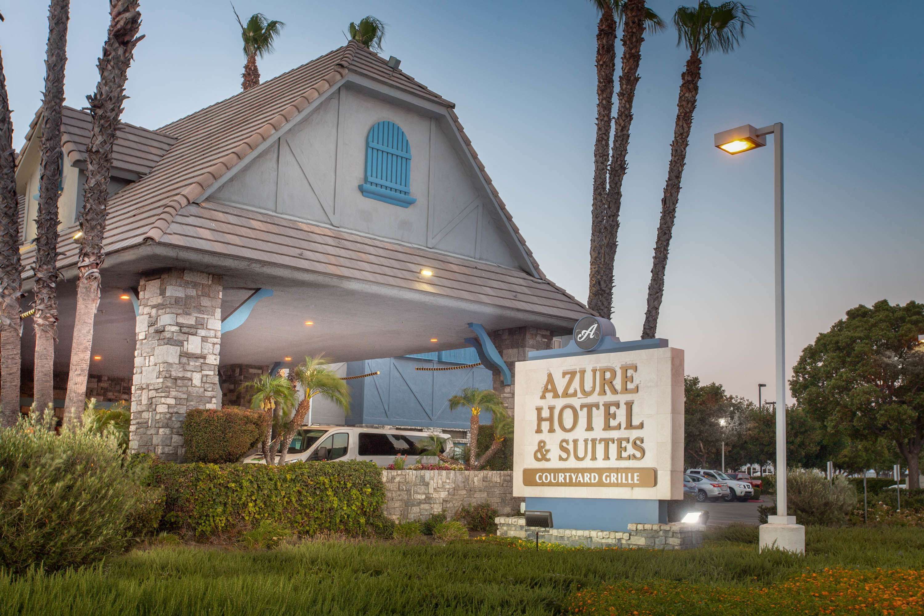 The exterior of Azure Hotel & Suites with a stone facade, blue window shutters, and a sign indicating its location in the Courtyard Village.