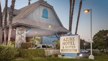 The exterior of Azure Hotel & Suites with a stone facade, blue window shutters, and a sign indicating its location in the Courtyard Village.