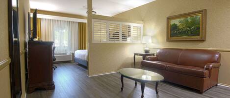 Une chambre d’hôtel avec un lit, un canapé en cuir marron, une table basse en verre, un bureau avec une lampe, un tableau au mur et une fenêtre avec des rideaux.