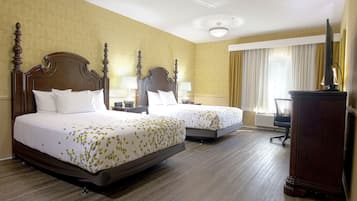 Habitación de hotel con dos camas, cabeceros de madera, un escritorio con silla, un televisor de pantalla plana y un ventanal con cortinas.